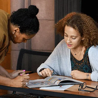 A student examines a book while her professor looks on.