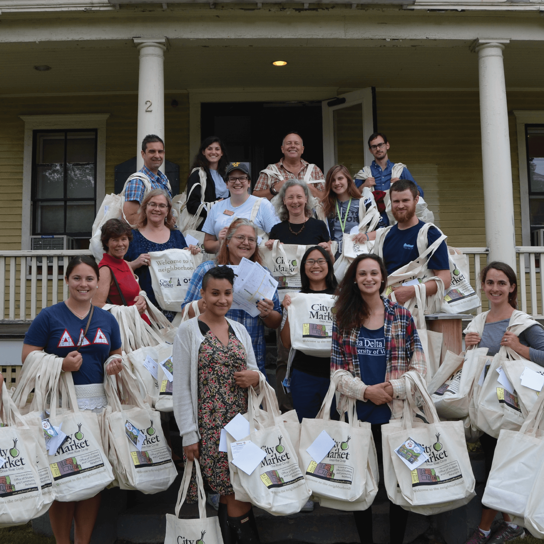 Group of volunteers holding reusable bags that read, "Welcome to the Neighborhood" stand on the porch steps of Pearl House.