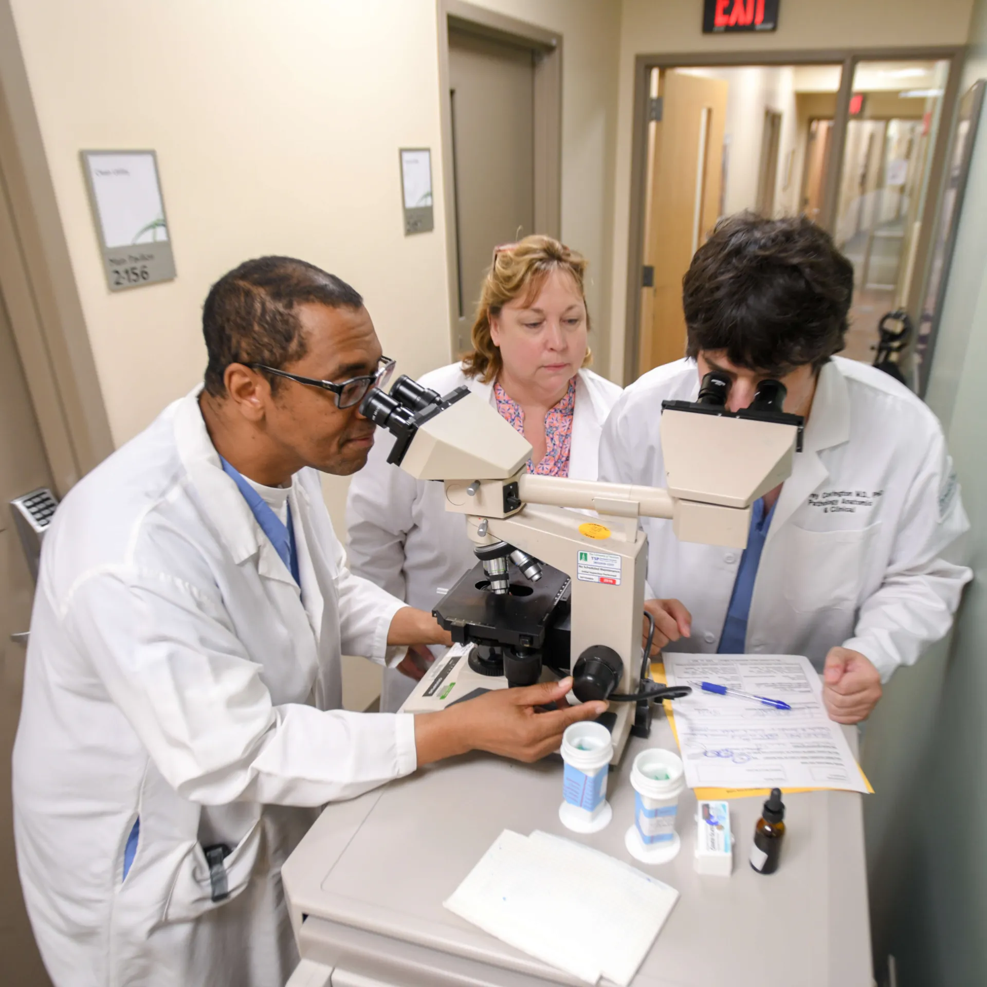 Three doctors standing at a microscope, two of them looking through the microscope lens.