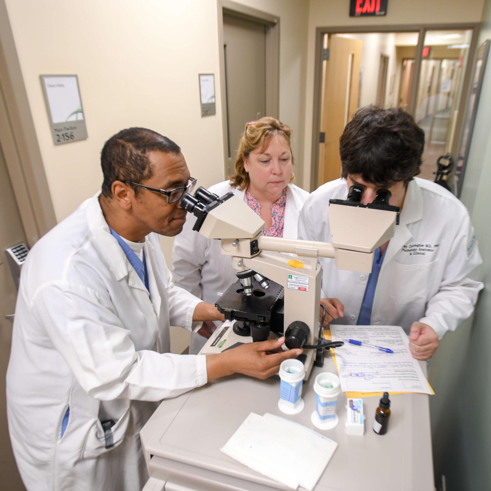 Three doctors standing at a microscope, two of them looking through the microscope lens.