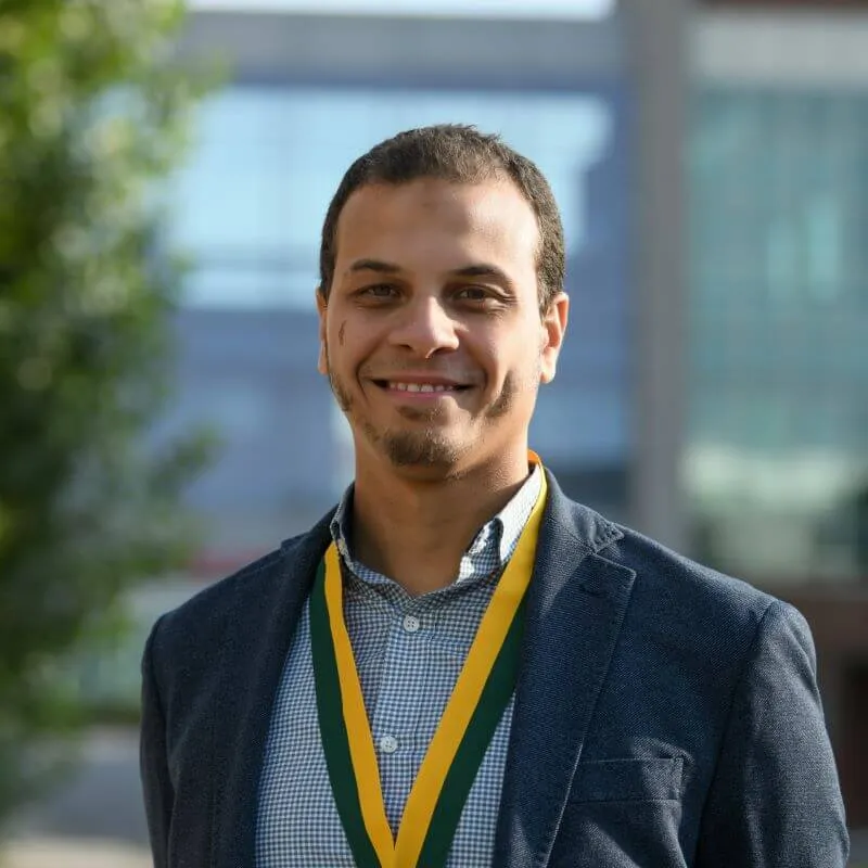 Headshot image of the researcher adorned with an award, posed outdoors in front of a distant building.