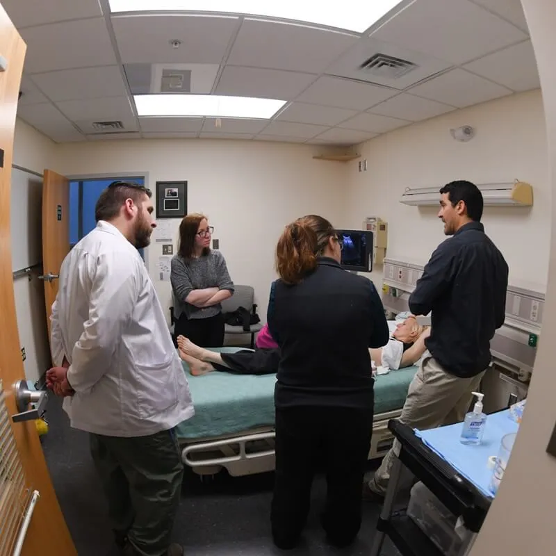 A group of learners gathered around a patient on a bed, listening to their instructor.