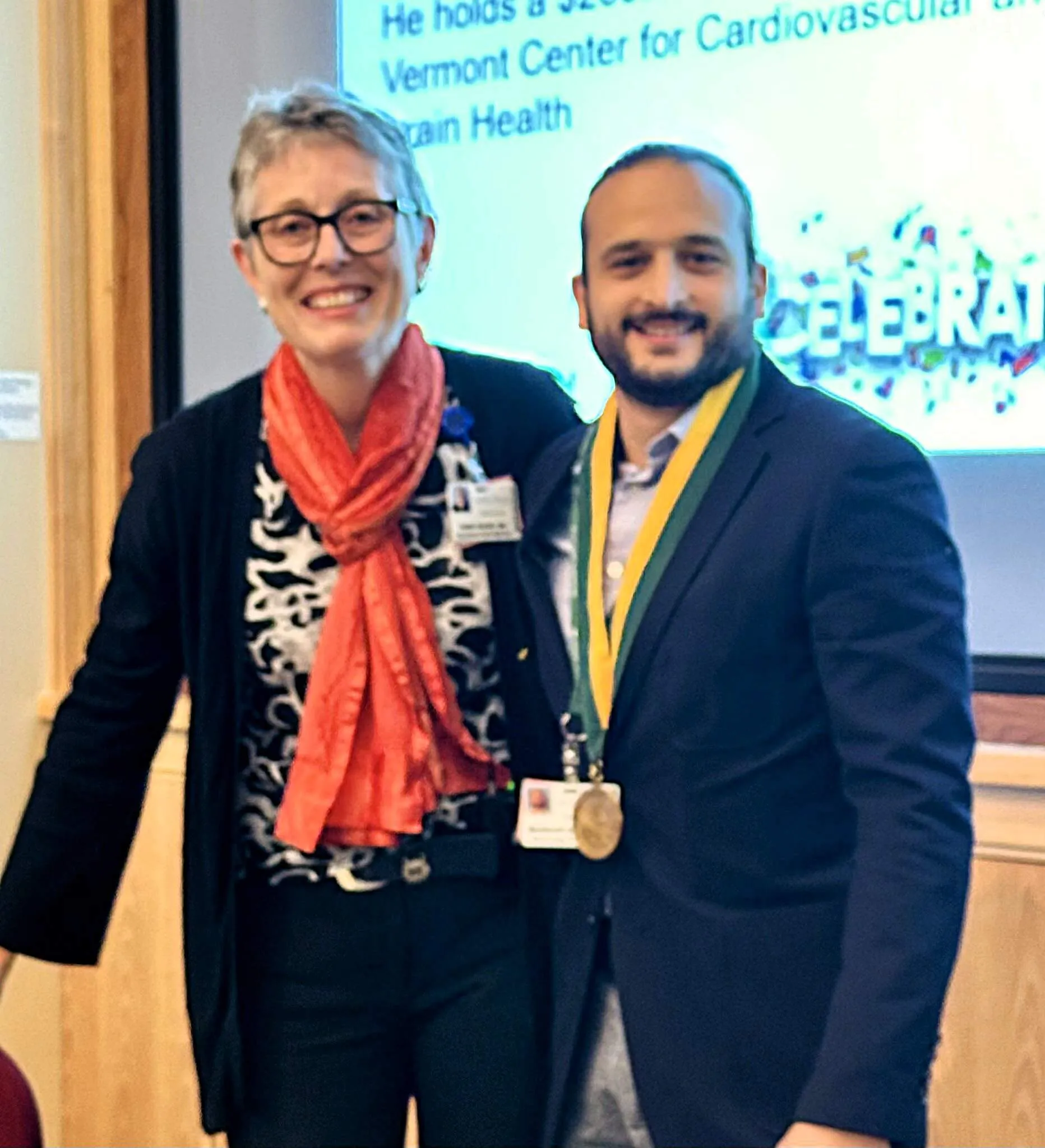 Two researchers are posing together, smiling, with one of them adorned with an award while posing indoors.