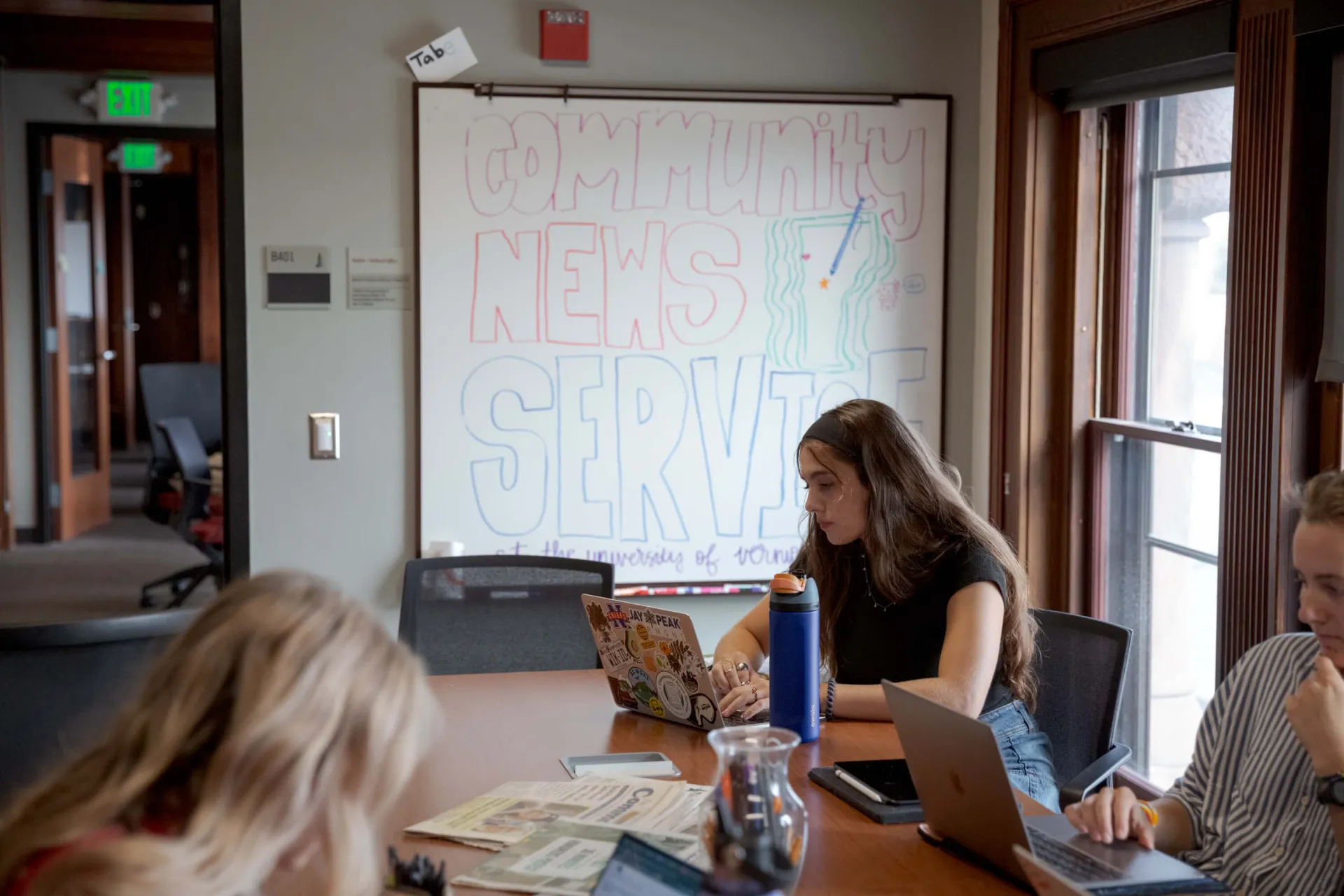 A University of Vermont student sits at a busy conference table working on her laptop. She is in front of a whiteboard that says "Community News Service."