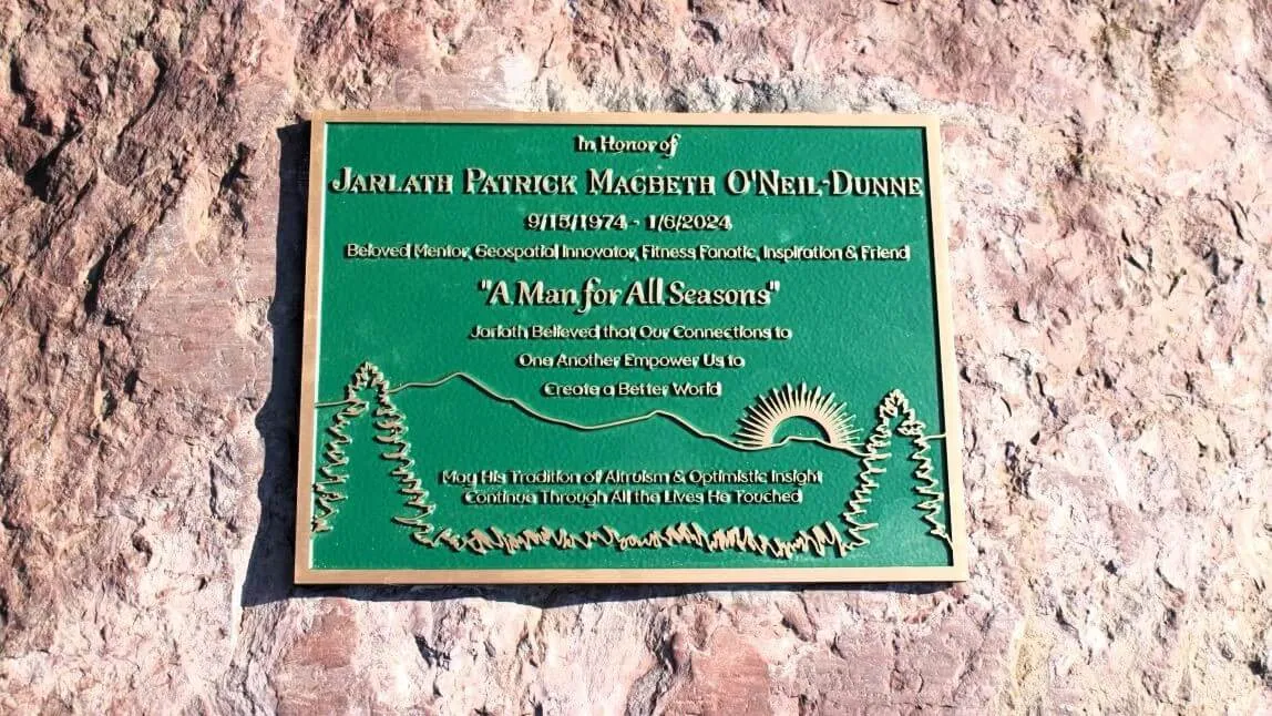 The memorial plaque for Jarlath O'Neil-Dunne placed outside the Aiken Center at UVM - the plaque is green and gold and depicts an enscription, trees, mountains, and the sun.