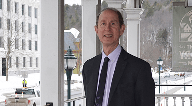 a man in a suit standing with the UVM campus in the background