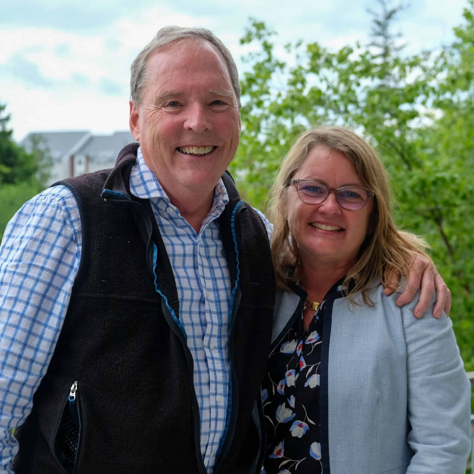 2 people smiling outside with green foliage and a blue sky in the background