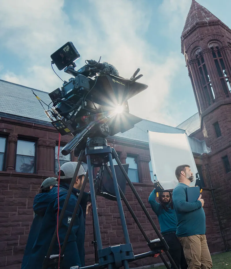 A group of people gathered around a camera in front of a building, engaged in conversation