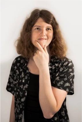 esmée in a black shirt and black and white shortsleeve button down in front of a white wall, thumb and forefinger on her chin