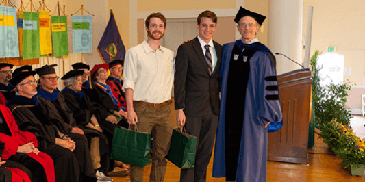 alt="two honors students with their faculty mentor on stage in full regalia"