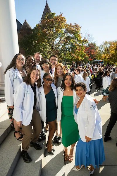 A group of smiling people standing outside on the steps of a building on a sunny day, wearing white coats. 