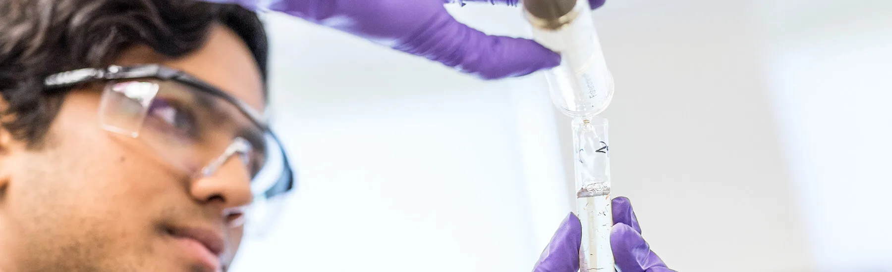 A person wearing a lab coat and purple gloves is holding a test tube