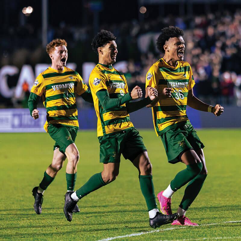 three students on a soccer field wearing uniforms cheer a win