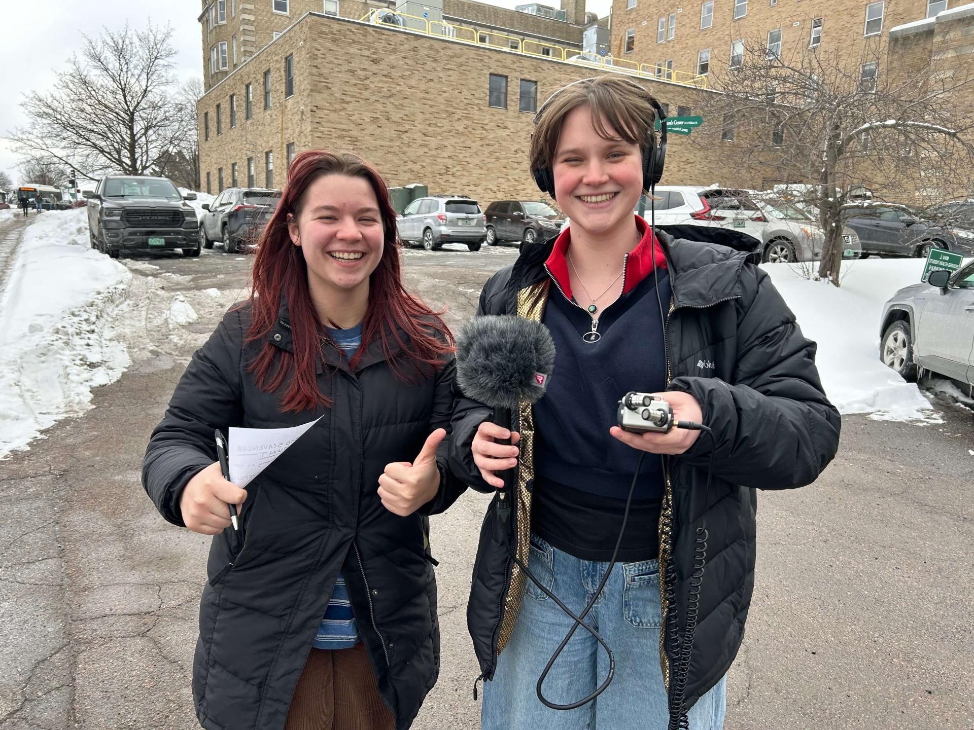 Two university of Vermont students wearing coats hold a microphone and notes while they cover the 2024 federal election.