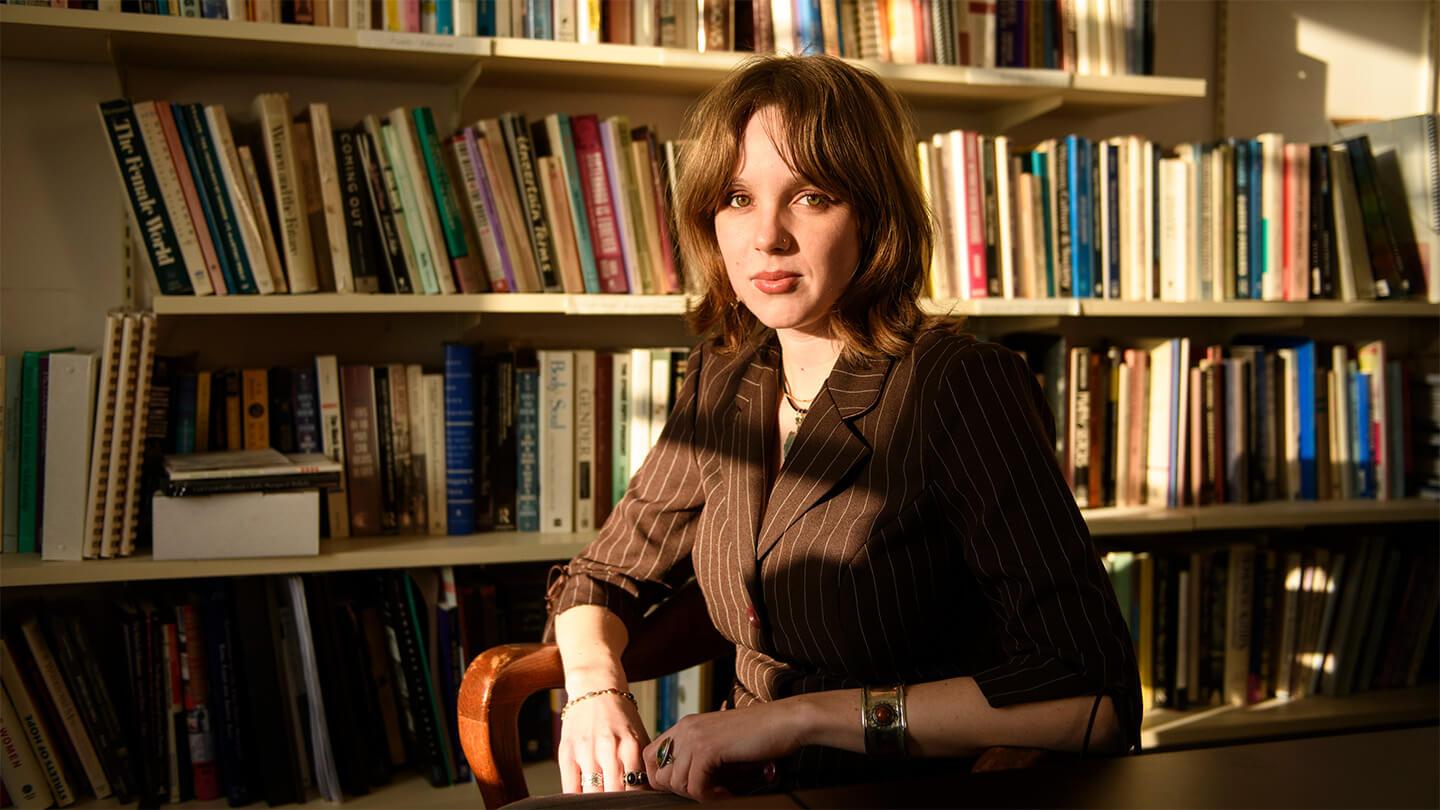 Young woman with brown hair wearing brown suit sitting at table in front of bookshelf