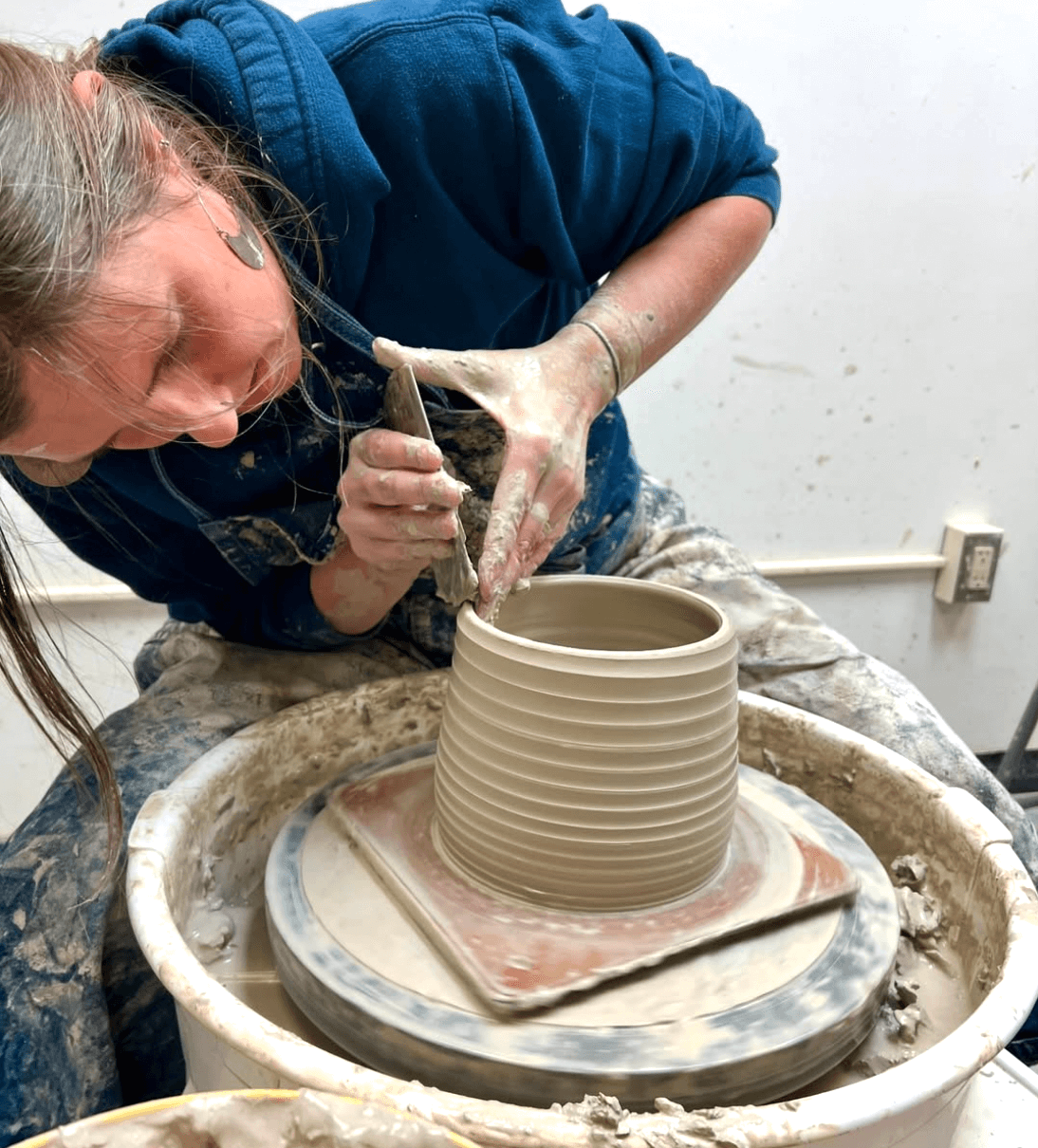 A person is focused on crafting a clay pot on a potter's wheel, surrounded by pottery tools and materials.
