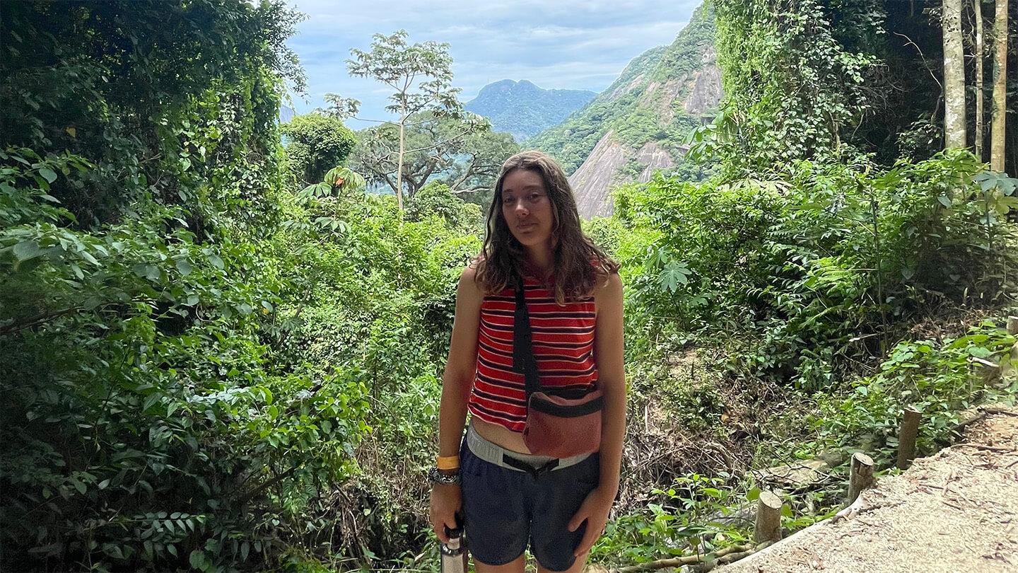 Young woman with long brown hair and orange top standing on path surrounded by greenery with mountain in distant background