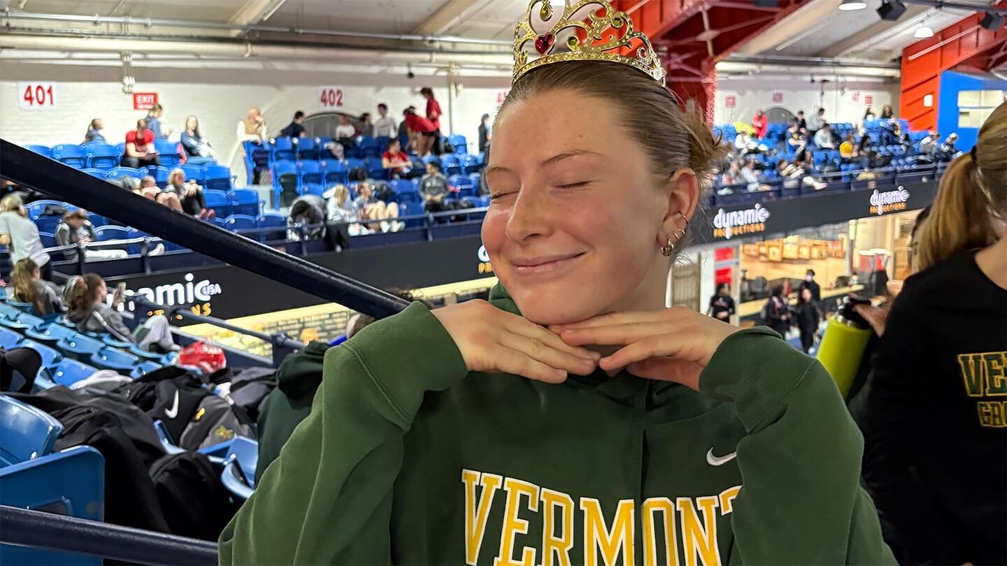 Young woman with brown hair pulled back in bun, wearing green UVM t-shirt with "Vermont" spelled out in yellow, standing in gym bleachers with eyes closed and hands under chin