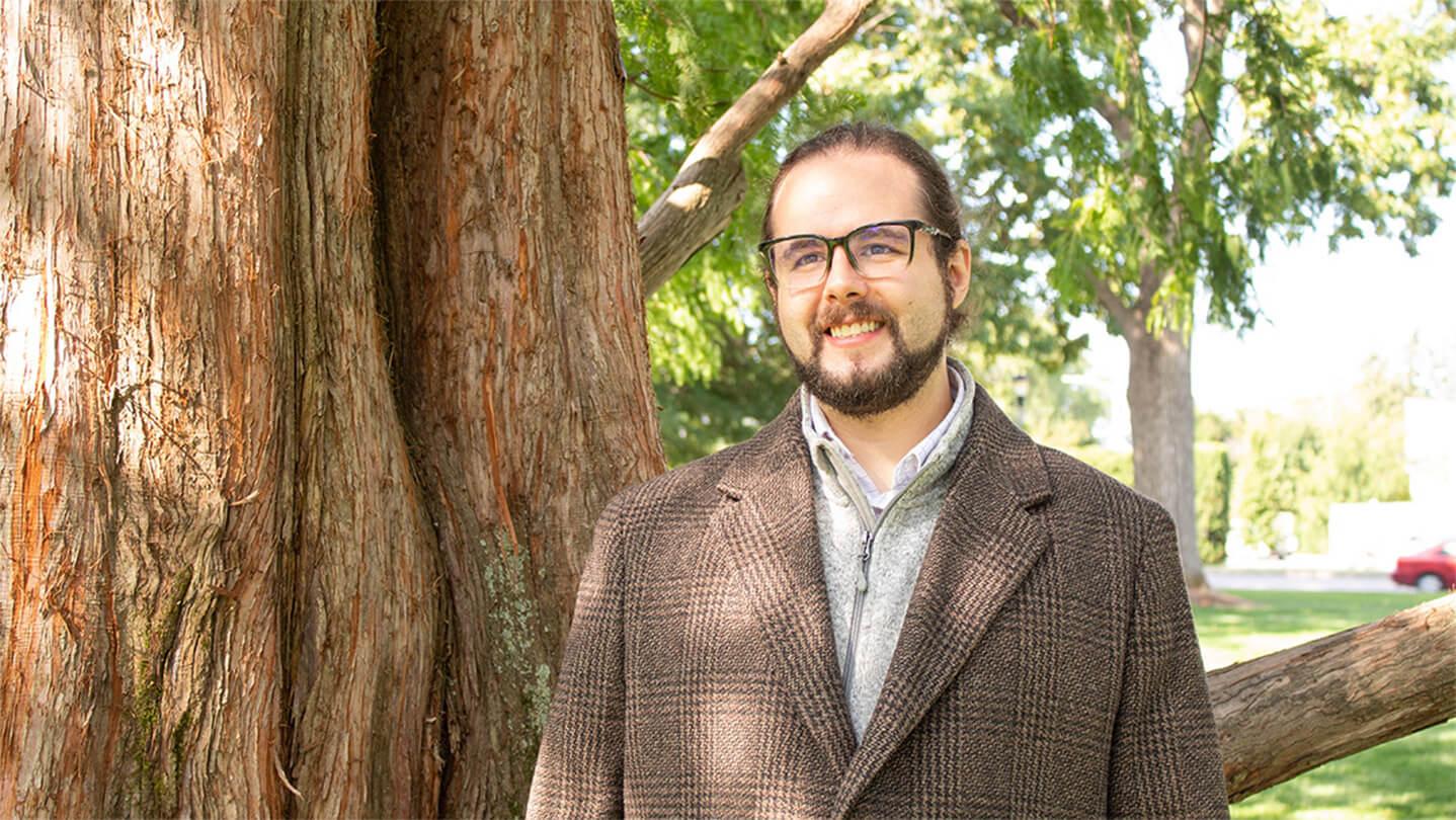Man with brown hair and beard, wearing glasses and a brown plaid coat, standing by a tree on UVM Green