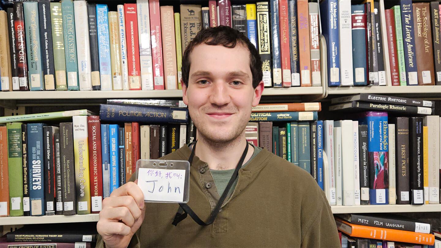 Young man with brown hair and green shirt in front of bookshelf holding name tag written in English and Chinese
