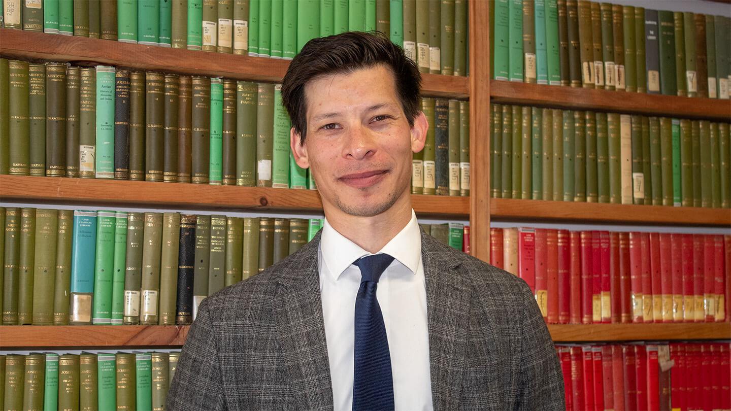 Young man with brown hair in plaid jacket with white shirt and dark tie standing in front of bookshelf with green and red books