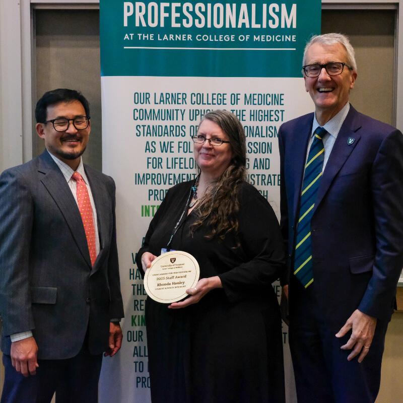 woman holding a plaque is flanked by two men in suits