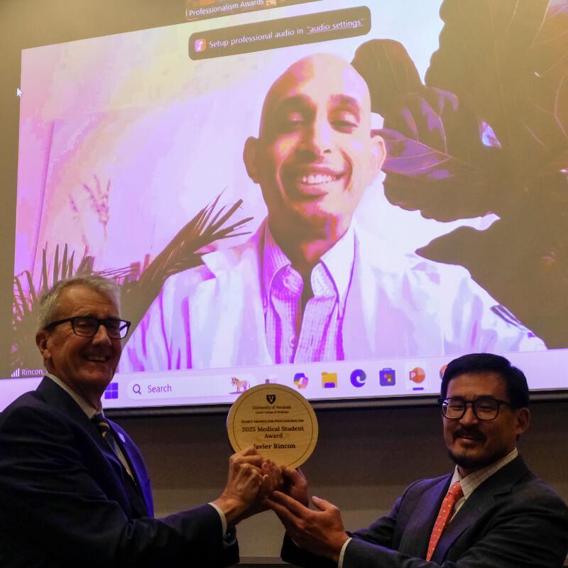 two men hold an award plaque while winner is on a video screen in the background