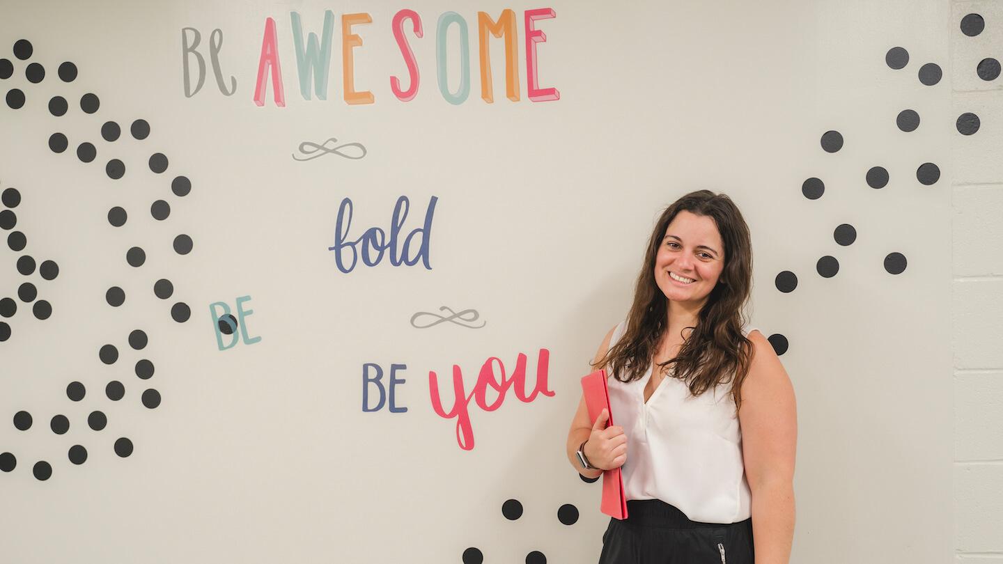 Monica Desrochers at the South Burlington School District in front a wall mural graphic with the words: "Be Awesome Be Bold Be You"