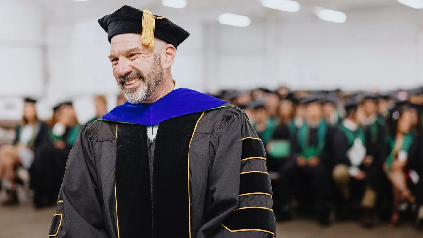 Peter Newman in his regalia in front of a crowd of graduating students that are blurred in the background, Peter is smiling