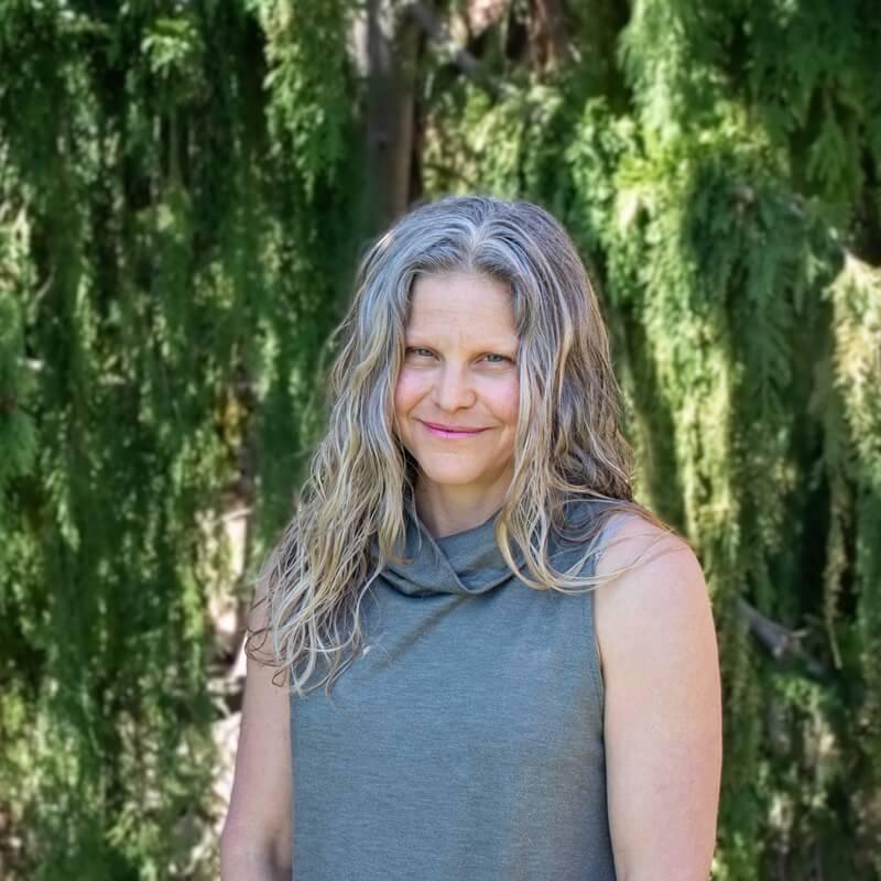 Rachelle Gould headshot, she is wearing a grey sleeveless shirt and is standing in front of a green, forested background