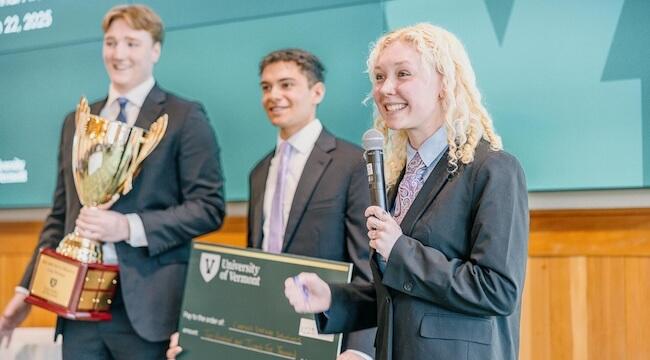 three students standing, one talks into microphone