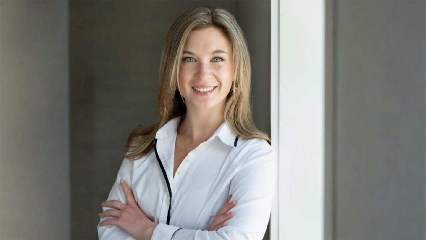 Professional headshot of young woman with long blond hair wearing white shirt; she is standing with arms crossed, smiling