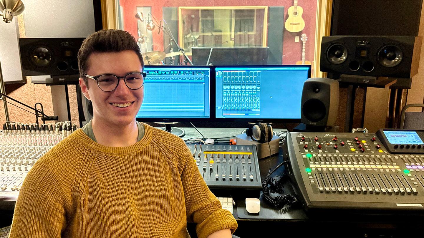 Young man with brown hair, glasses, and yellow sweater sitting in front of sound boards in music studio