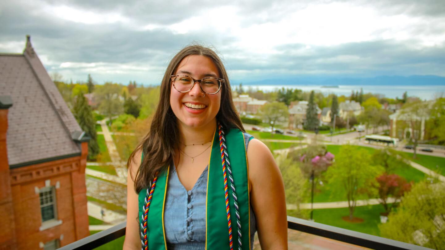 Isabel Wolhandler dressed in regalia on top of Old Mill building with the university green, Burlington, and Lake Champlain in the background.