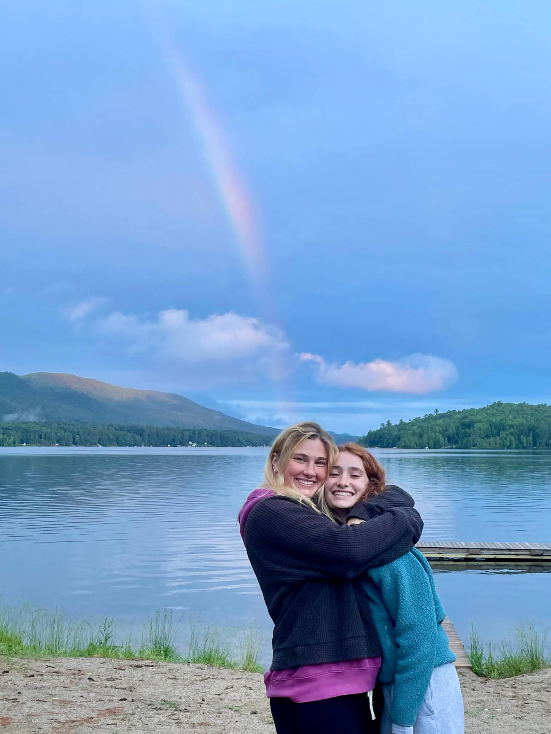 Chloe and a friend in front of a lake with a rainbow in the sky.