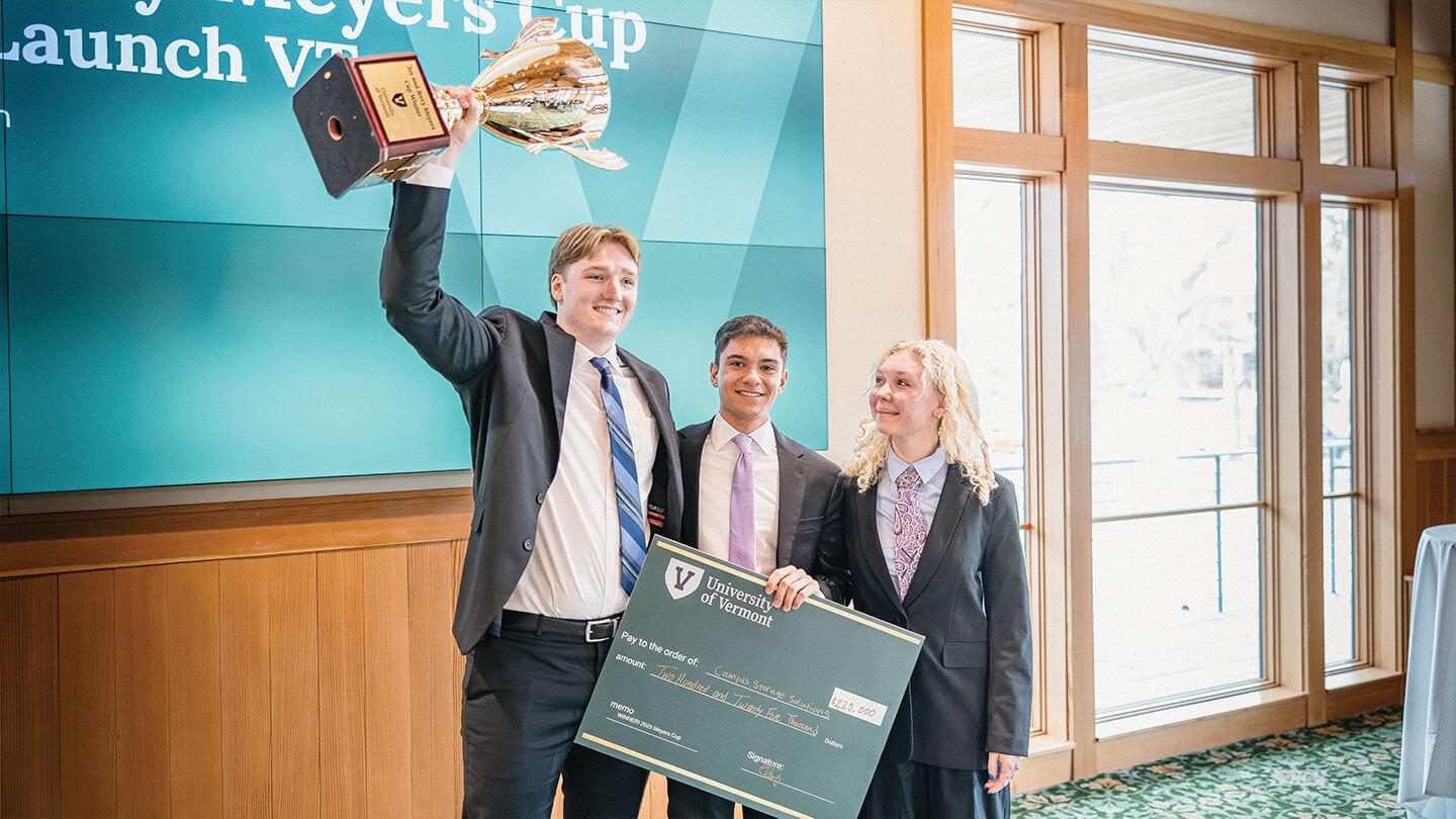 three students cheer while holding a green oversized check and a trophy