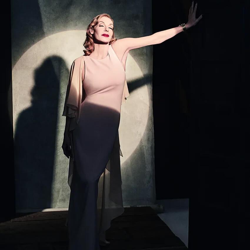 Ute Lemper standing in a spotlight wearing a pale pink dress