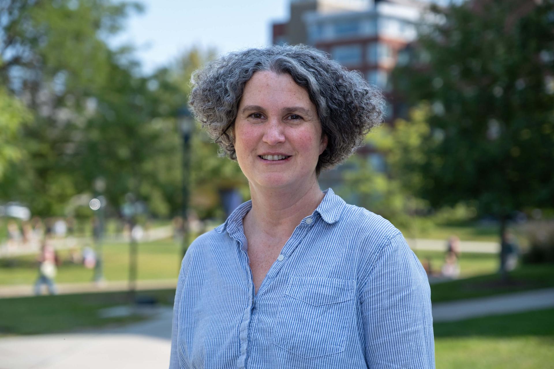 A woman in a blue button down shirt and short, gray curly hair stands on a college quad.