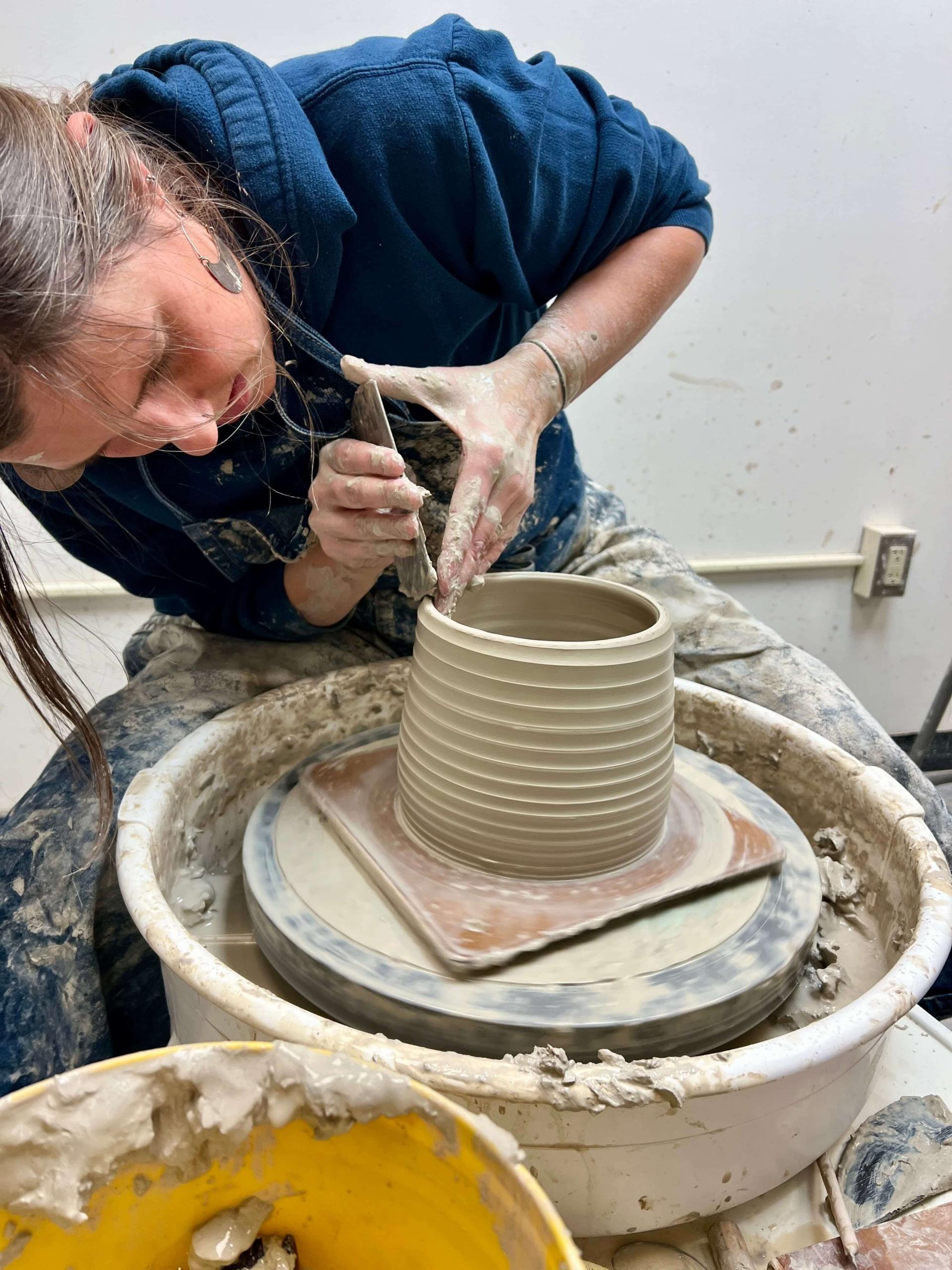 A person is focused on crafting a clay pot on a potter's wheel, surrounded by pottery tools and materials.