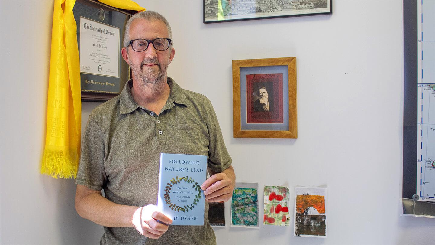 Male professor wearing beige polo shirt with goatee and glasses standing by wall in office holding new book