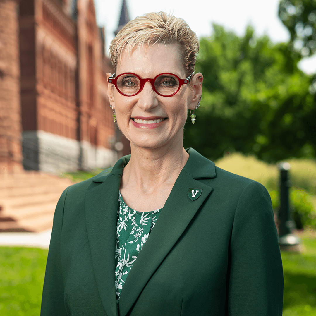 a person wearing red glasses and a green blazer poses in front of red brick buildings