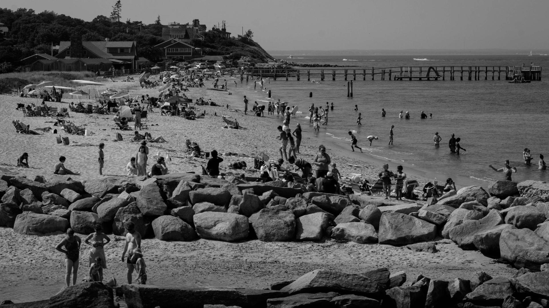 a black and white photograph of Martha's Vineyard, a beach with lots of people standing on it. There is a rocky jetty in the foreground