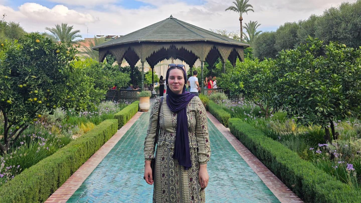Young woman in patterned sari and dark purple headscarf standing in front of tiled walkway leading to a Moroccan pavilion