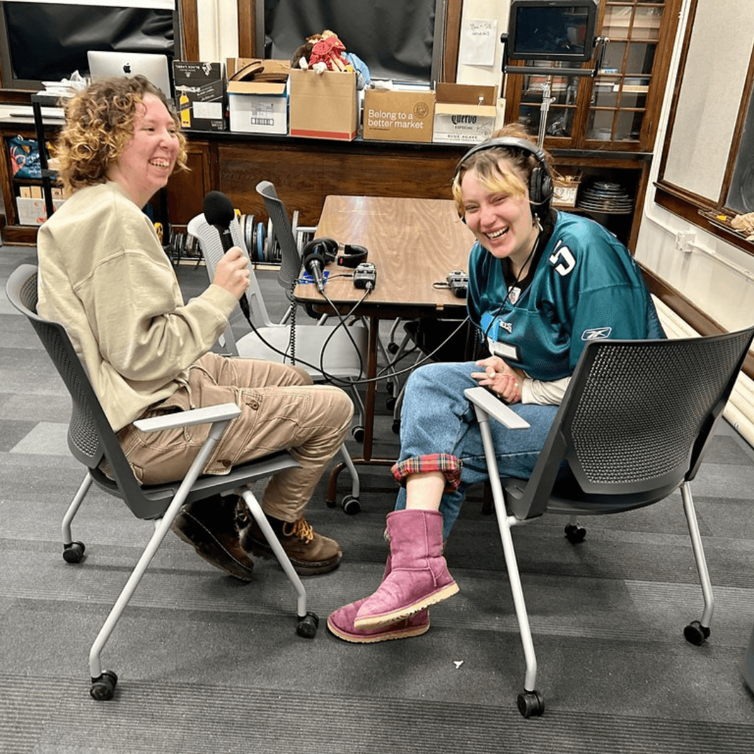 Two students in the UVM public radio class laugh together as they record with microphones and headphones.