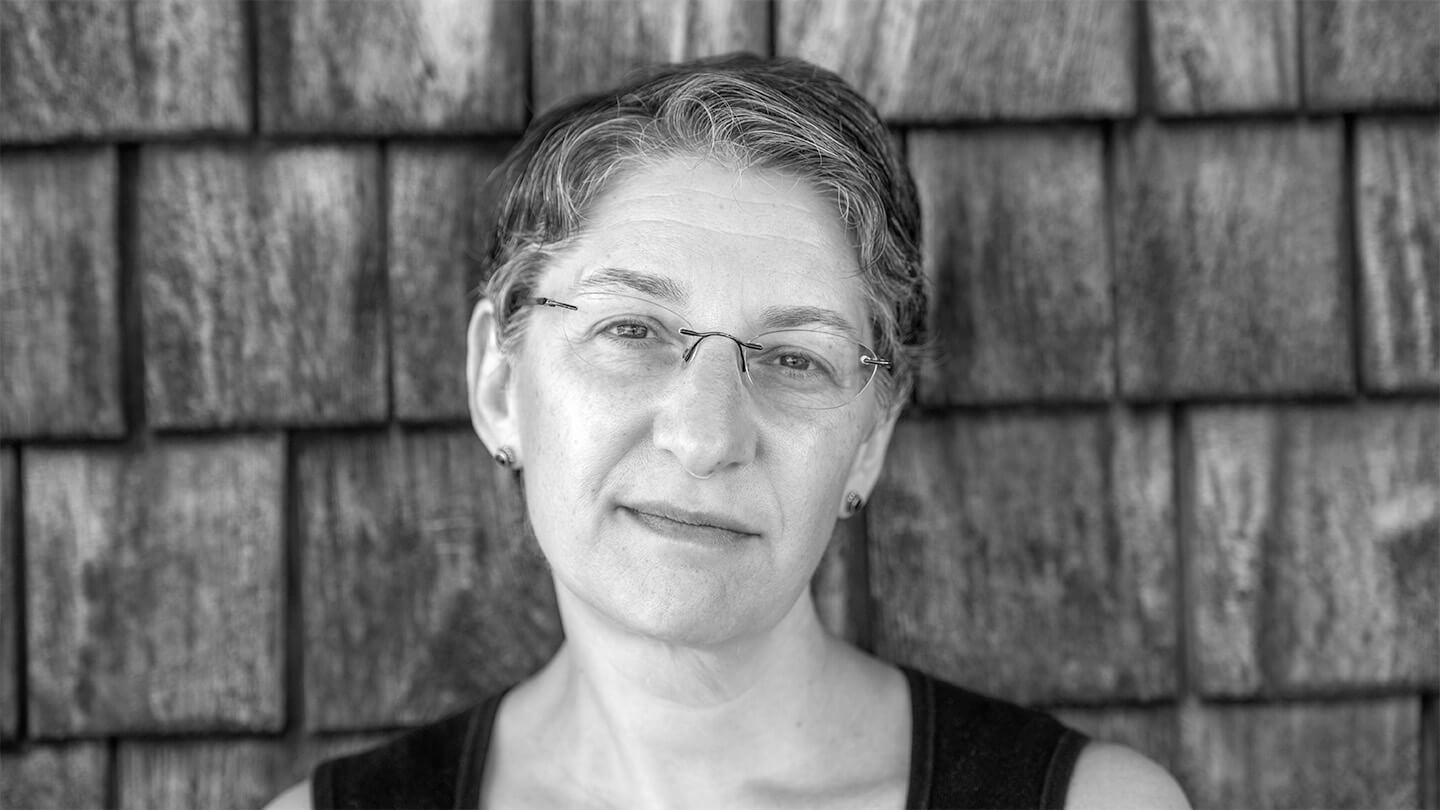 woman with short hair and glasses in front of shingled house, looking at camera (in black and white)