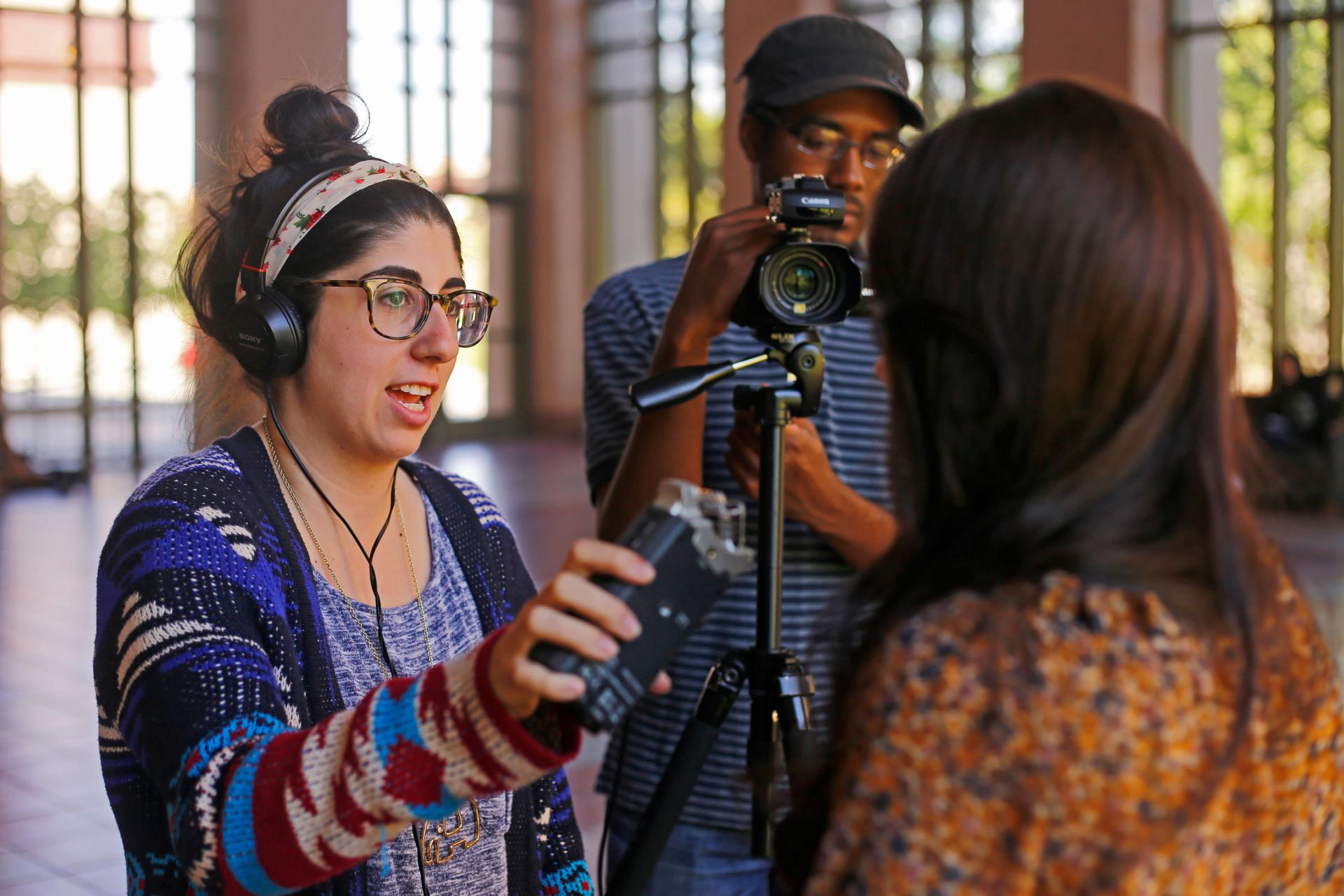 Student reporters Elvin and Renee interview a community member. Renee holds a microphone, wearing headphones and a colorful sweater.