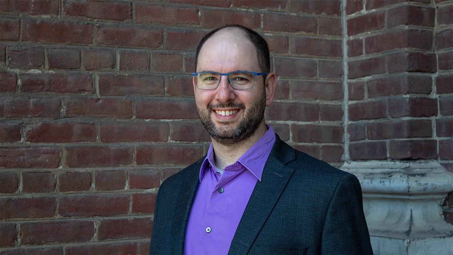 man with glasses and beard, wearing purple shirt and gray jacket, smiling and standing in front of a brick building