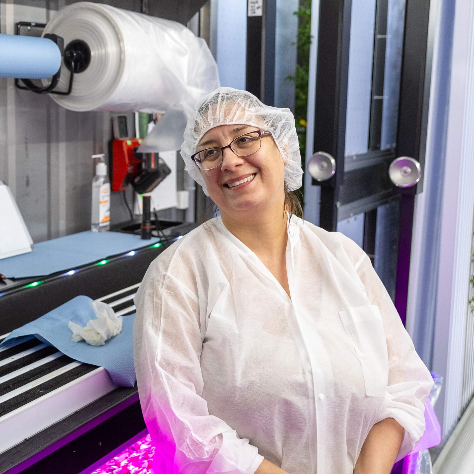 Worker in a hairnet and bodysuit smiling in front of seedling production area and vertical growing walls.