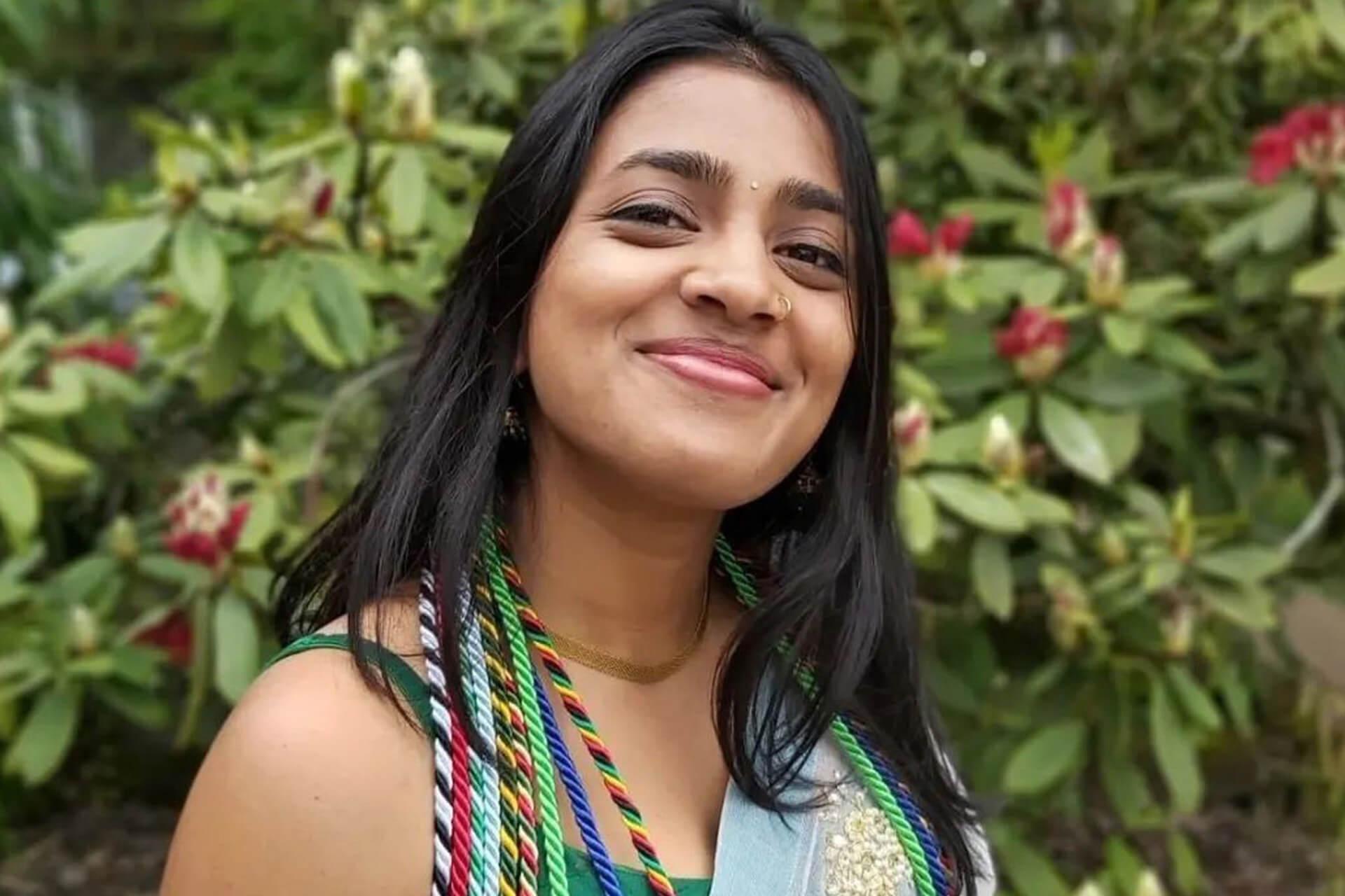 A person with long dark hair smiles at the camera, wearing graduation cords around their neck. The background is a green bush with red flowers.
