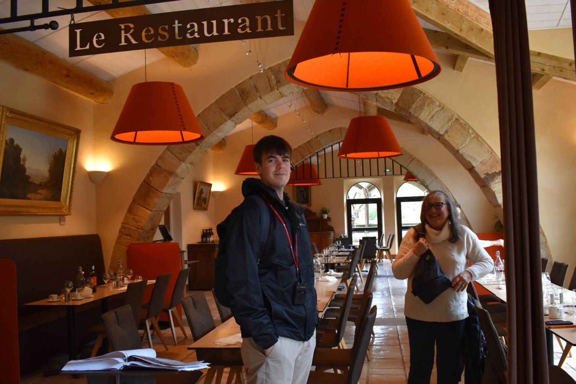  A person in a black jacket and a woman in a white sweater stand inside a restaurant with stone arched ceilings and a sign that reads "Le Restaurant."
